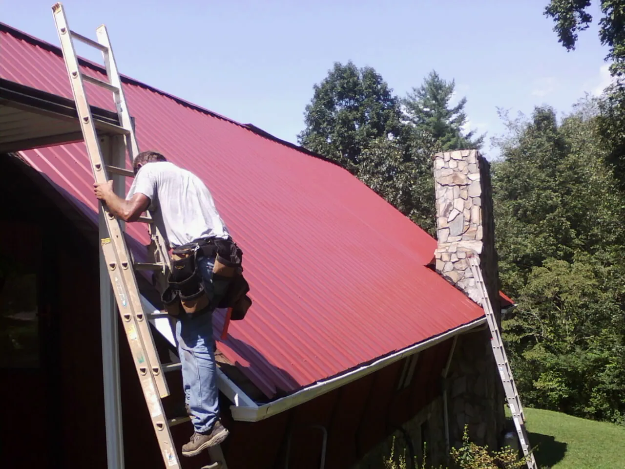 Roofer on a ladder beside a red metal roof and stone chimney — Soffit & Fascia Repair by Roofing Brothers in Fort Klamath